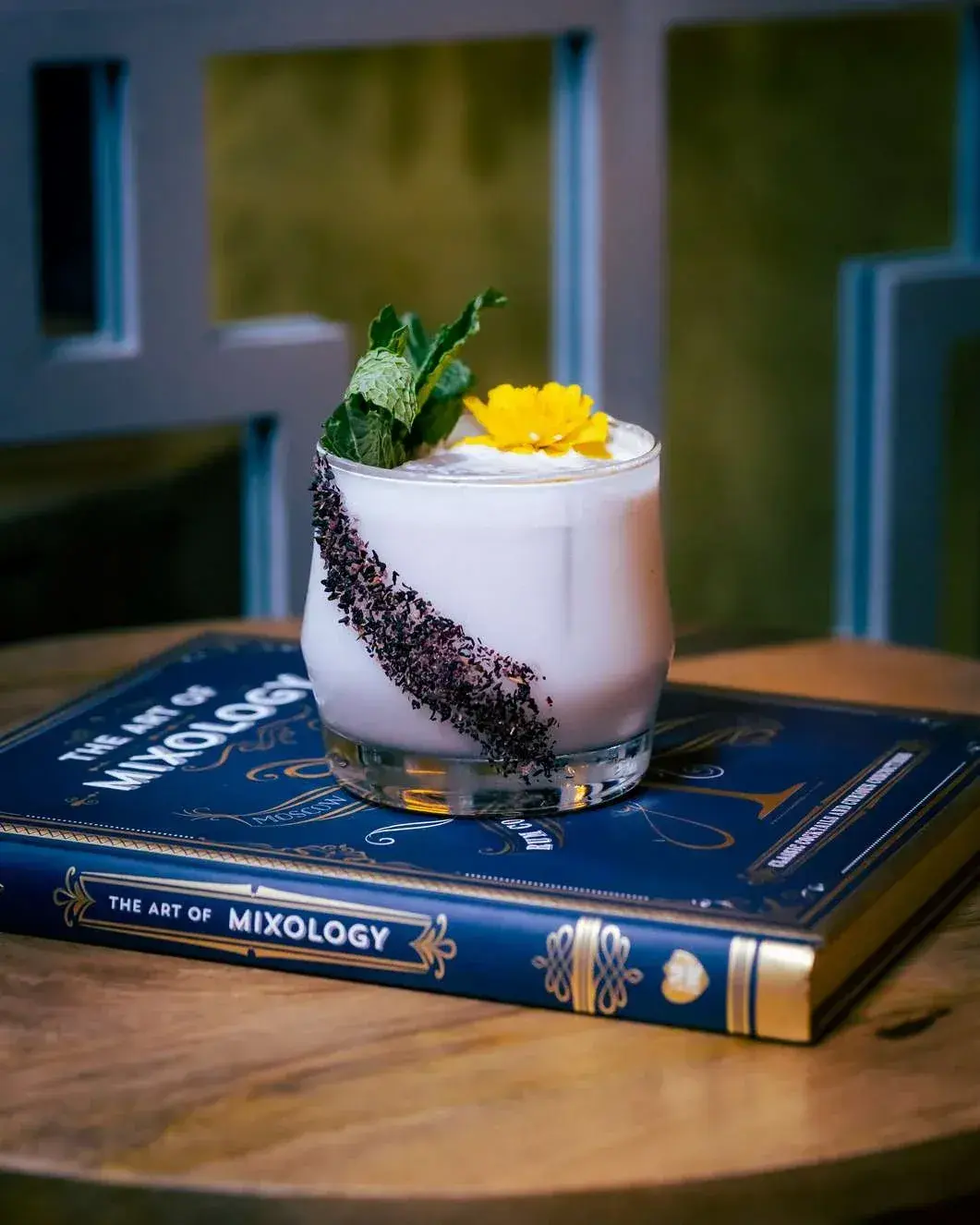 Creamy cocktail with mint and a yellow bloom, balanced on a mixology book