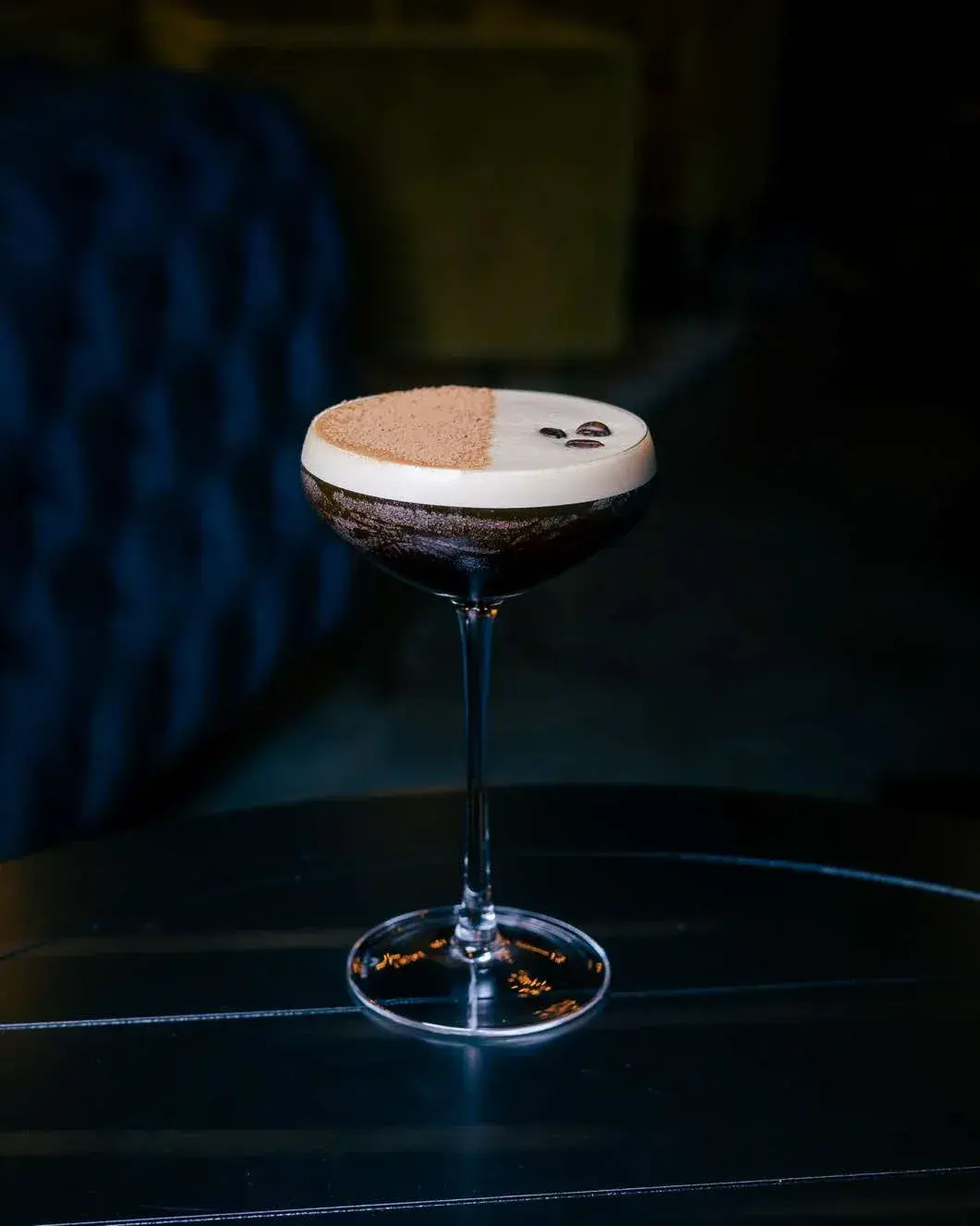 Espresso martini with cocoa dust and three coffee beans beside a blue tufted booth